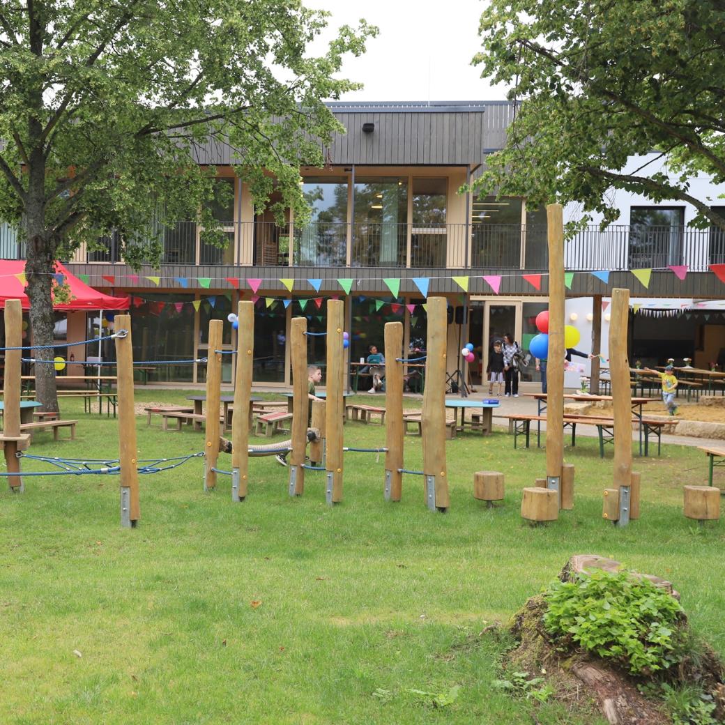 Man sieht das Außengelände mit einem Spielplatz und der KiTa im Hintergrund. Es ist eine bunte Wimpelkette gespannt.