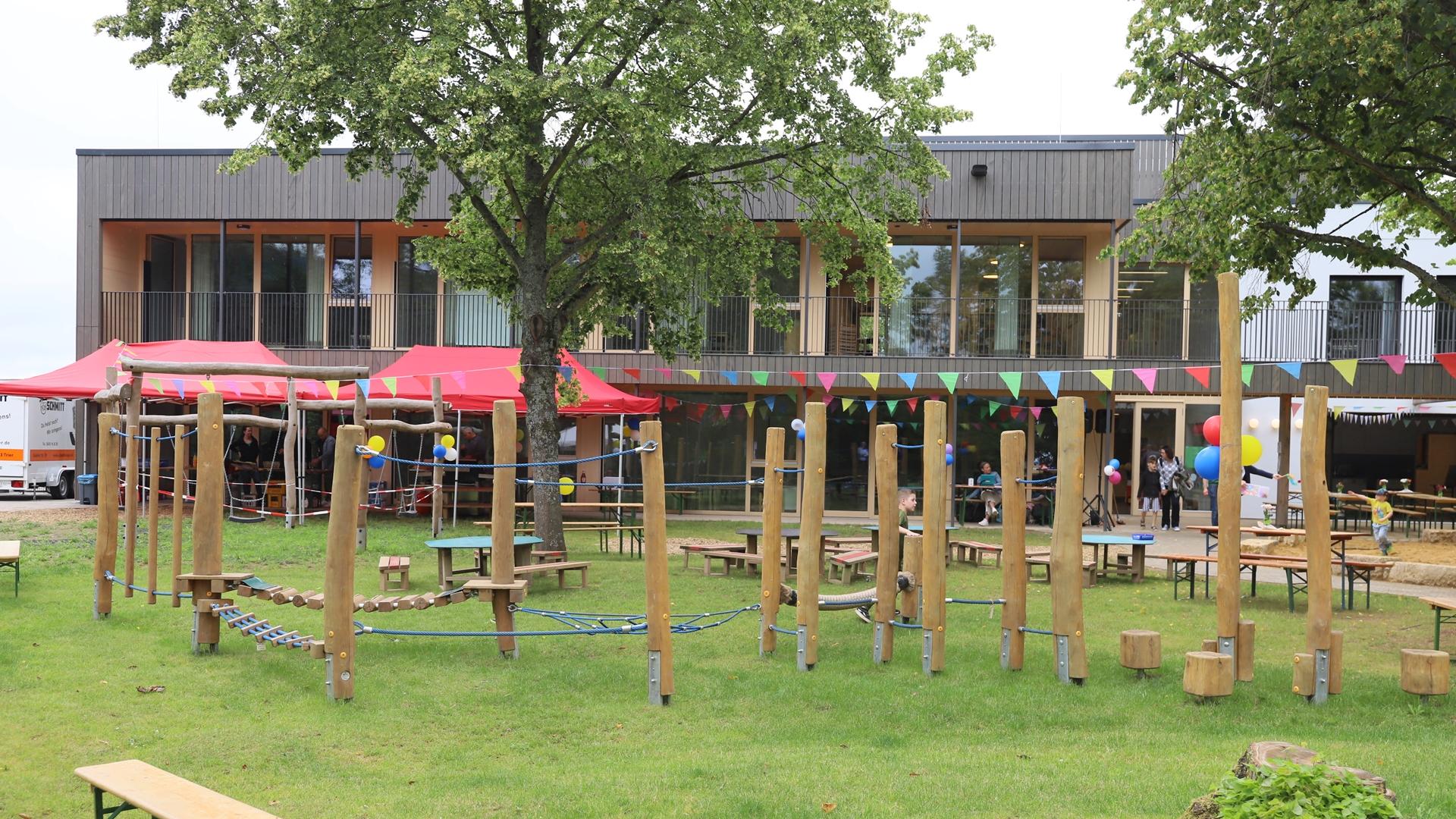 Man sieht das Außengelände mit einem Spielplatz und der KiTa im Hintergrund. Es ist eine bunte Wimpelkette gespannt.